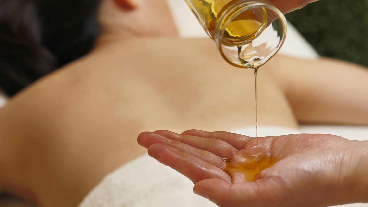 Spa staff pouring massage oil from glass jar in hand over woman lying on table
