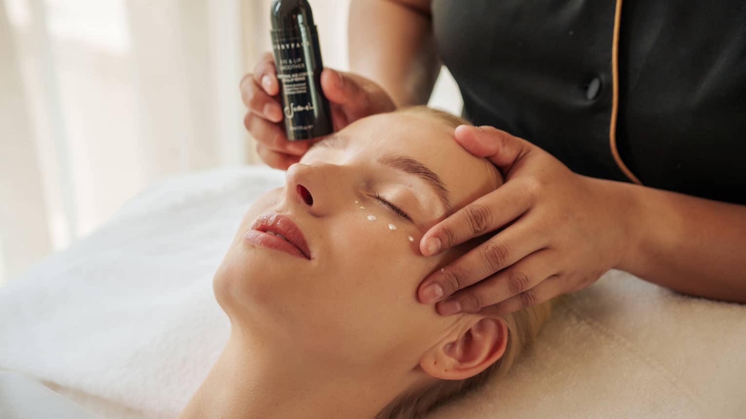Guest lies with eyes closed as a spa attendant places product under their eyes