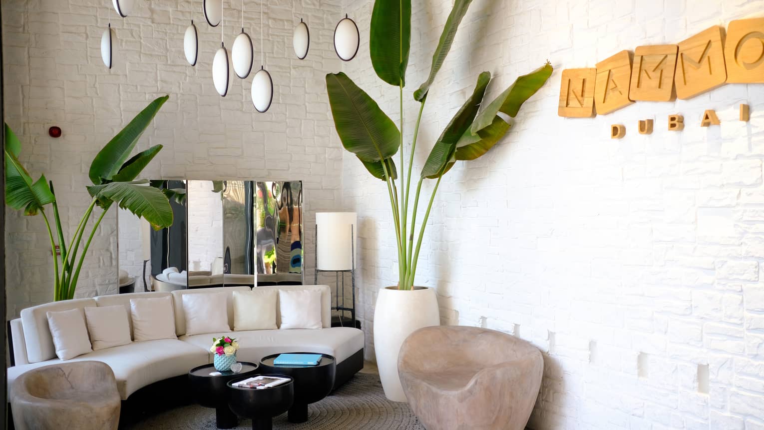 Indoor sitting area with curved white sofa, large palm plant, NAMMOS Dubai sign
