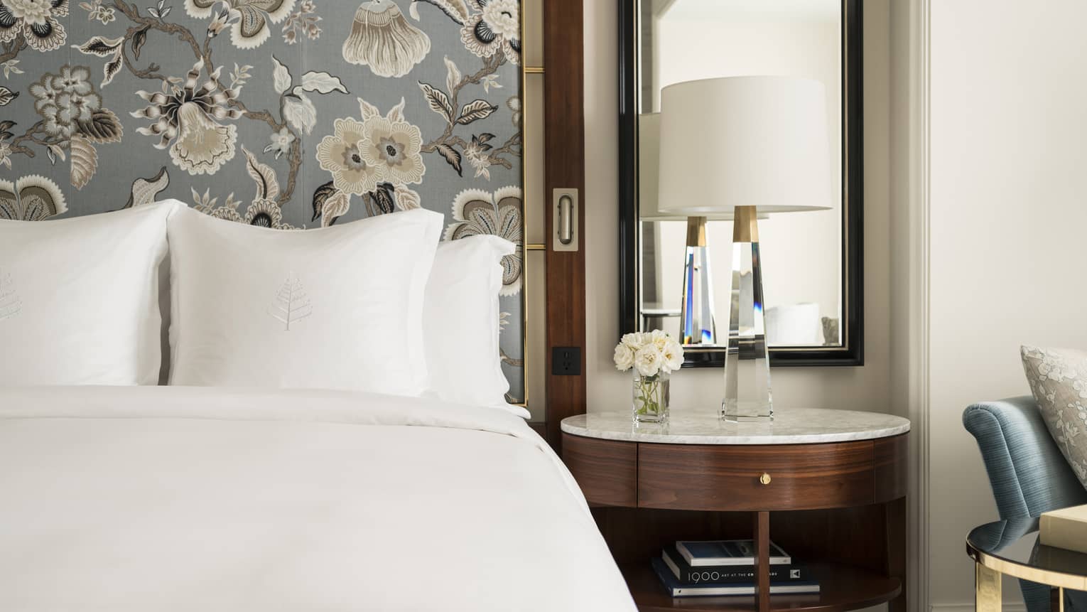 Close up of suite with white bedding, floral wallpaper, flowers, glass lamp on wooden table