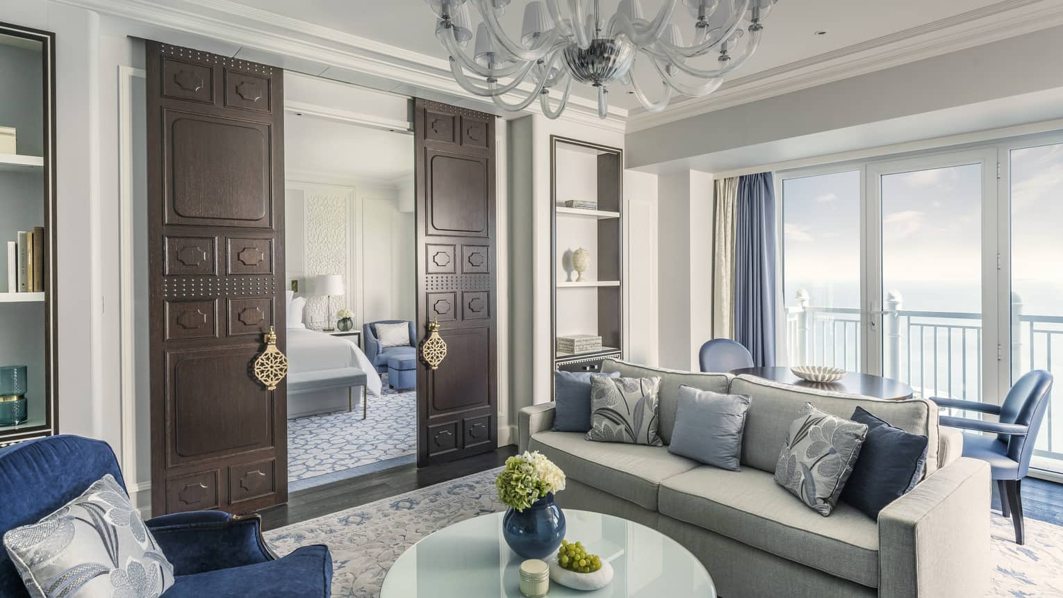 Sea-view living room, with grey sofa, blue pillows and accent chairs, chandelier, dark wooden doors open to bedroom