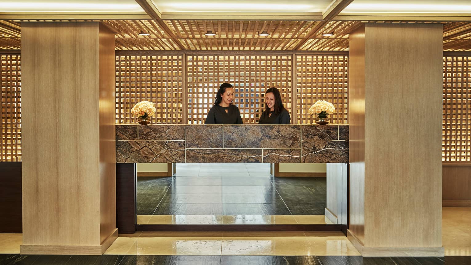 Two smiling hotel staff behind lobby front desk, wood pillars at side, wood lattice walls