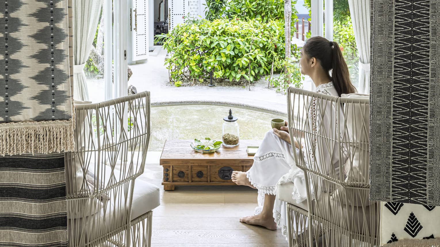 Person seated in a woven chair by a wooden table, enjoying tea in a serene spa waiting area with garden view