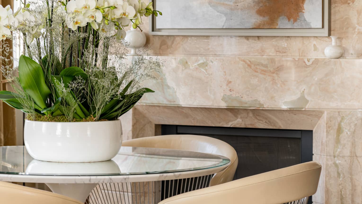 Close-up of a round glass-top table with beige chairs and a white orchid arrangement, set in front of a marble fireplace with wall art above