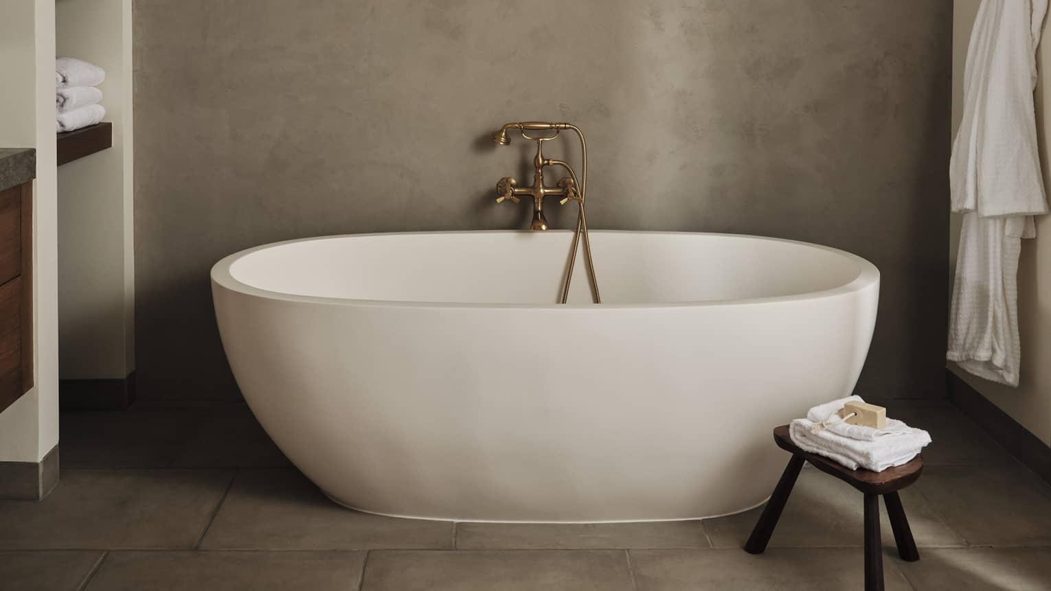 Luxury resort bathroom with a freestanding oval bathtub, built-in towel shelves, small wooden stool with folded towels and soap and bathrobe hanging on the wall