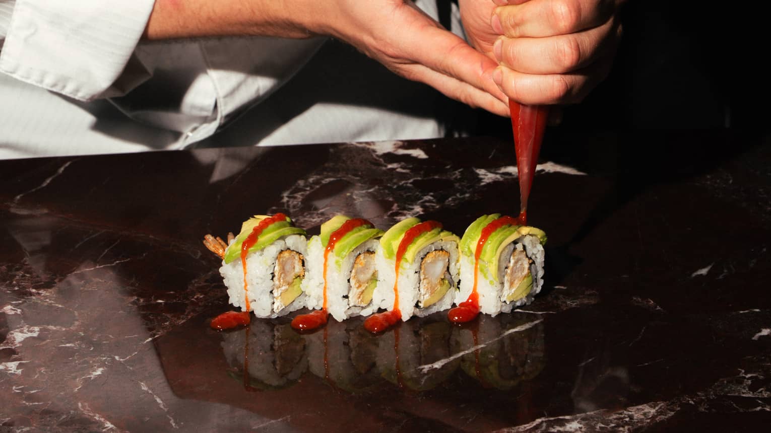 A chef adds garnish to a row of sushi rolls.