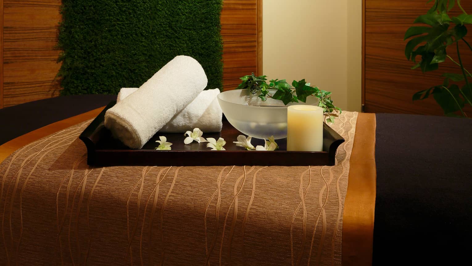 Wood tray with rolled white towels, flowers, candle, bowl with vine on spa bed