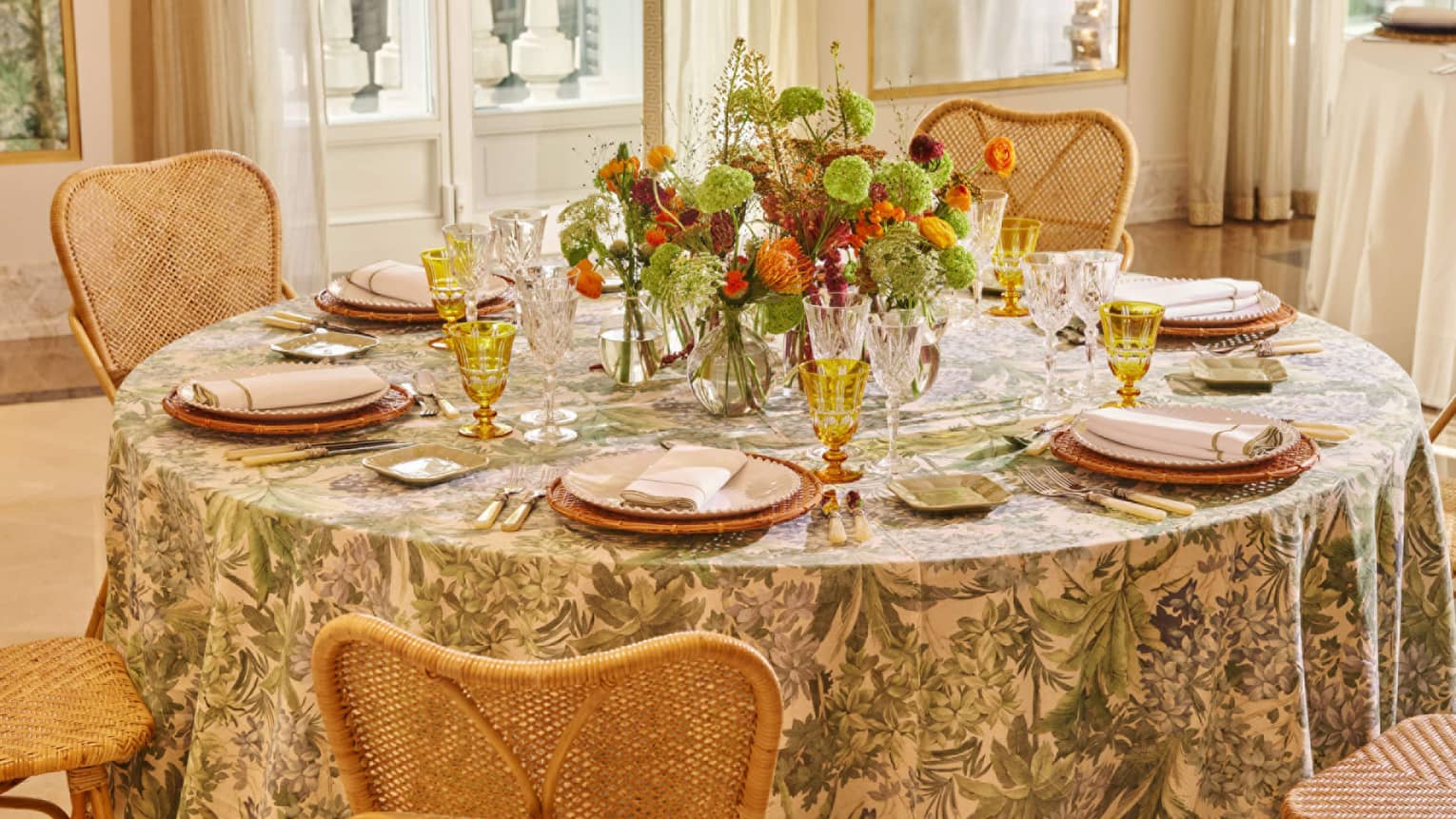 A well-lit interior event space with neutral decor features a round table with chairs and a floral tablecloth. The table is set with plates, napkins, utensils, clear and translucent gold stemware, small square side plates, and a colourful floral centerpiece.