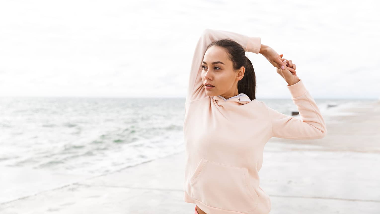 A woman in a pink hoodie stretching on a beach.
