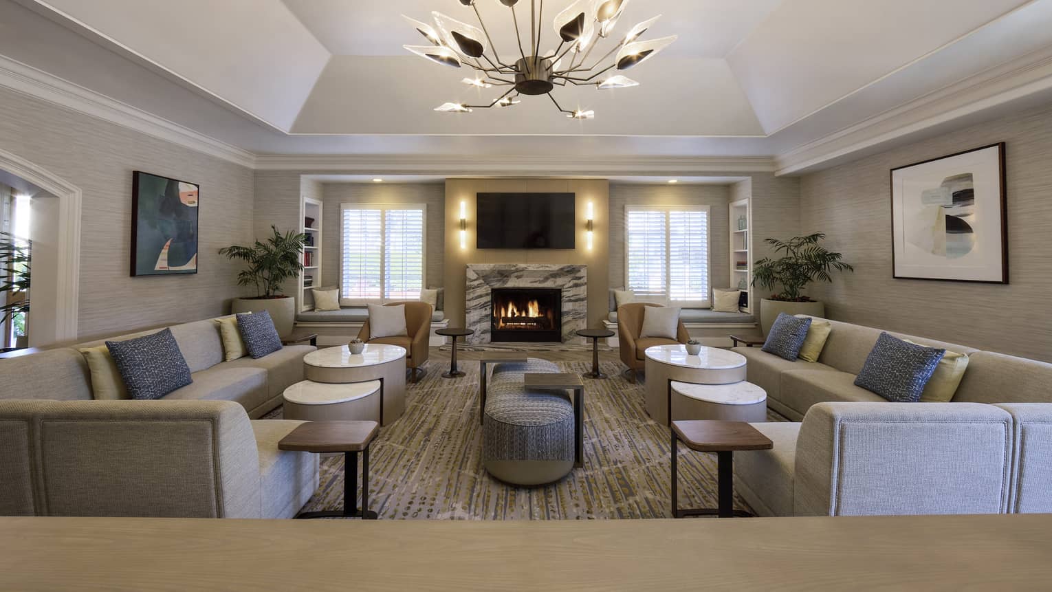 A spacious and inviting room with neutral decor and an artistic chandelier features couches with pillows, low tables, art on the walls, a fireplace, large potted plants, a flat-screen TV and windows admitting natural light.