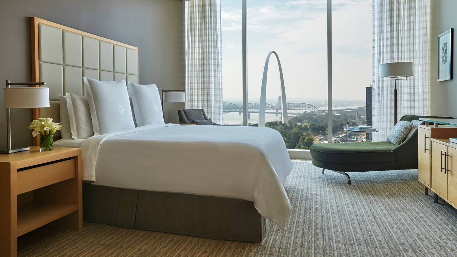 Bed with tall, padded white headboard, green velvet chaise beside floor-to-ceiling window with arch view