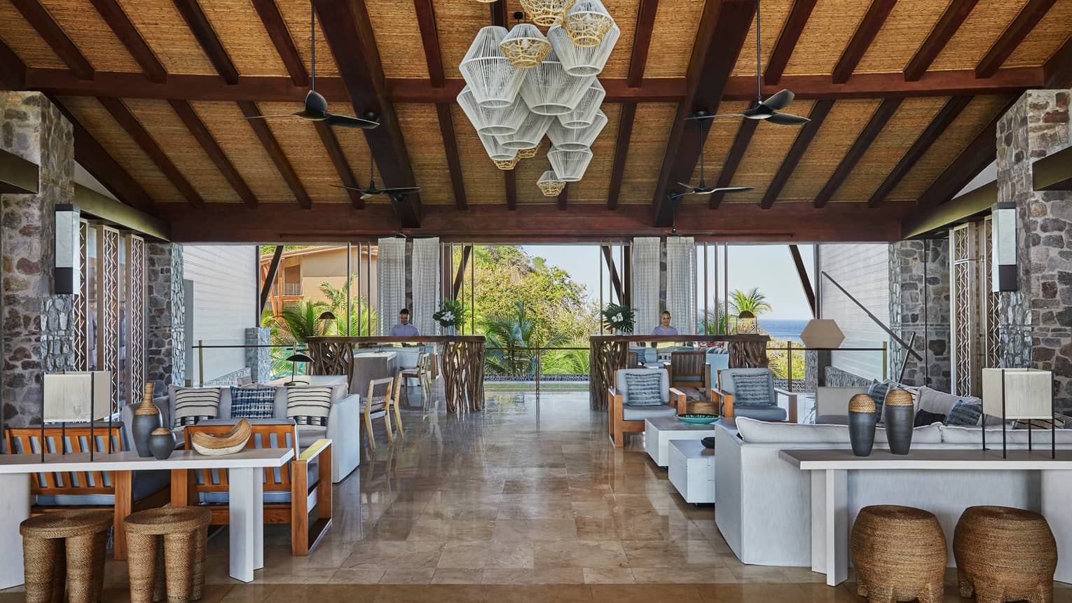Small sofas, tables with vases, rustic wood stools under modern wire chandeliers, rattan roof, in sunny lobby