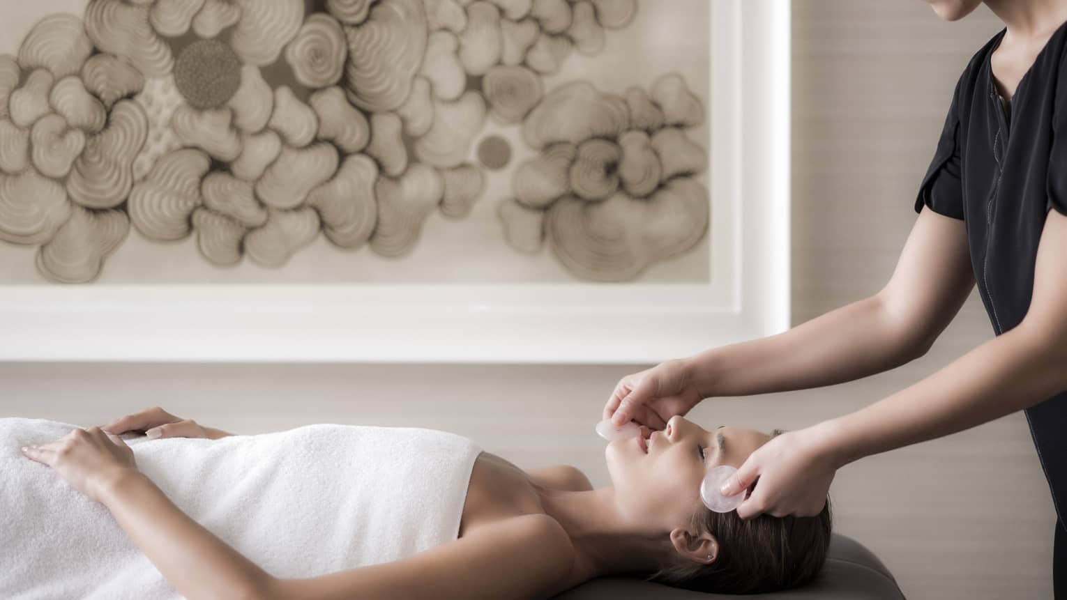 Woman lies on massage table wearing white towel as spa attendant massages her face with stones