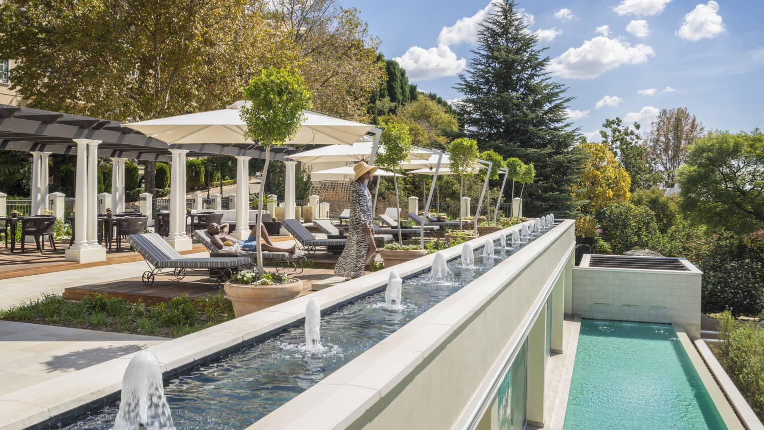 Main lounges on patio chair, woman stands by long fountain and ledge over plunge pool