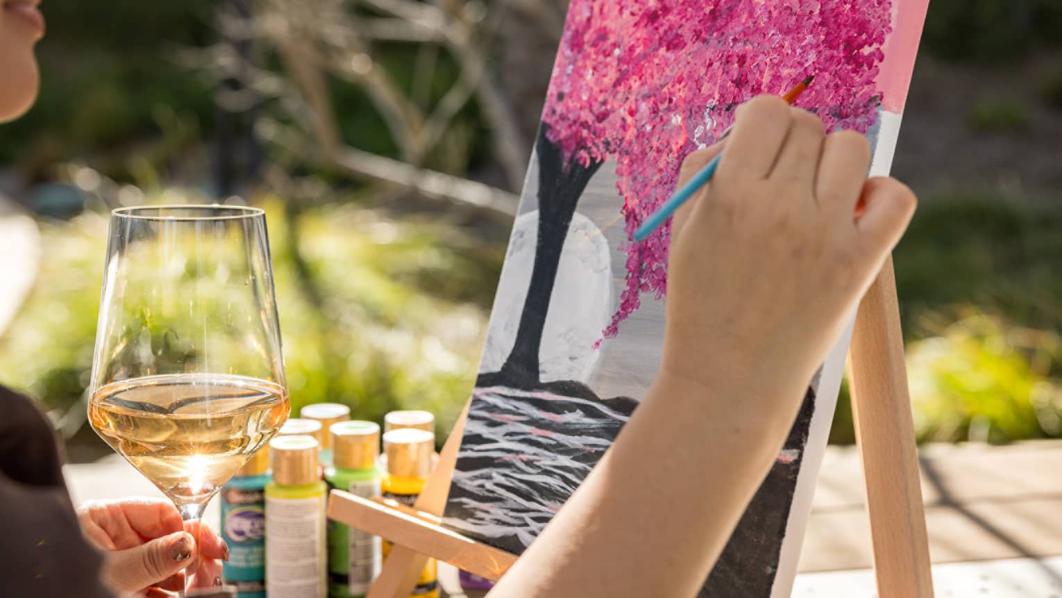 Person painting a pink tree on a canvas with one hand while holding a glass of wine in their other hand