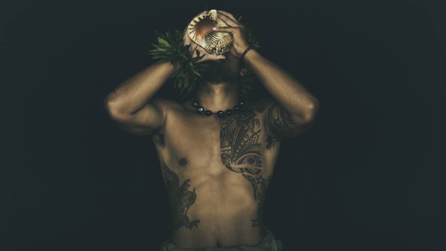 Shirtless tattooed Kānaka Maoli man plays traditional conch shell like a trumpet in the night