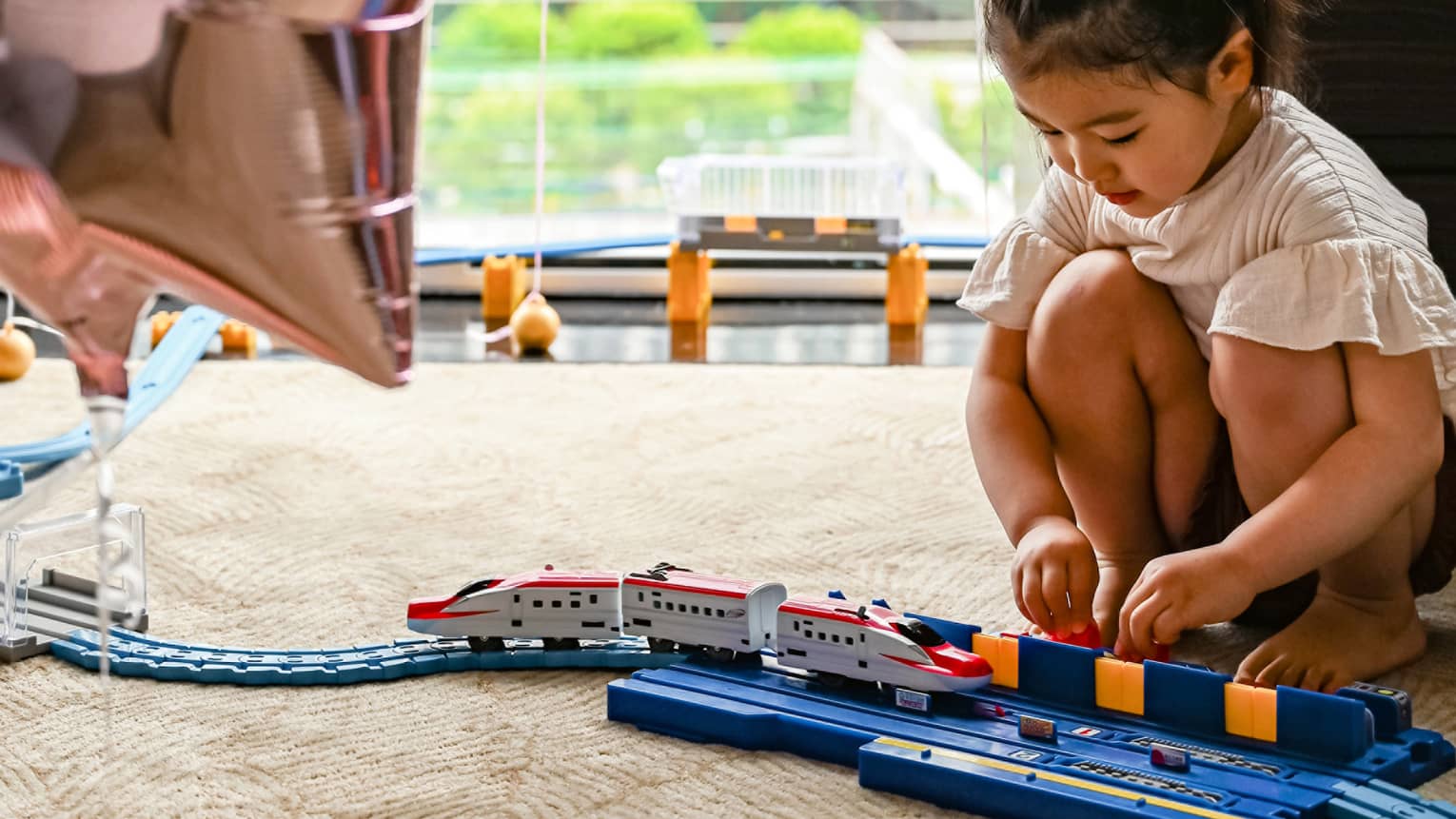 Young girl plays with train, large window and star balloons in the background