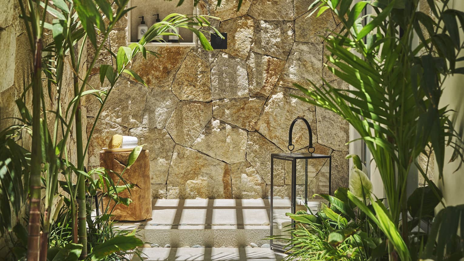 Casita outdoor shower area with a stone wall, surrounded by lush green plants, a wooden stool with towels and a decorative lantern