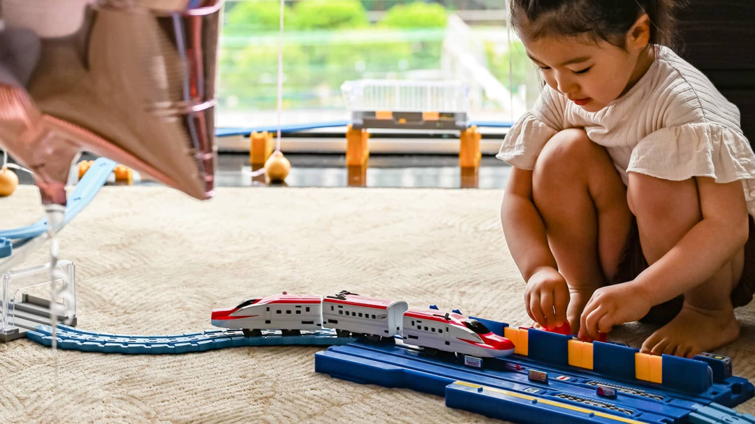 Young girl plays with train, large window and star balloons in the background