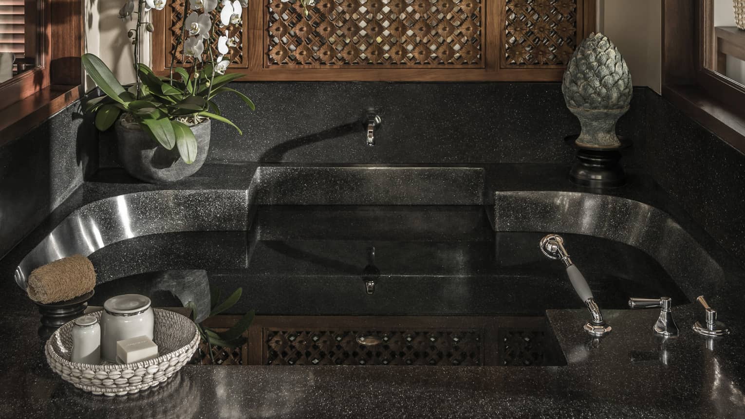 Black stone bathroom tub with potted plant and sculpture on ledge, basket with soaps and jars 