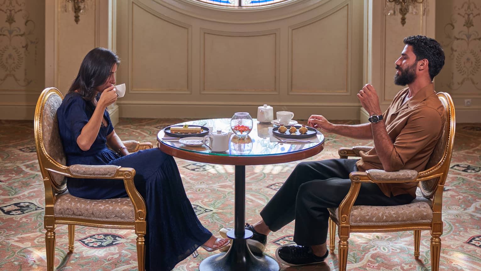 Couple sits across from each other sipping tea at small round table in front of large round window adorned with blues and greens