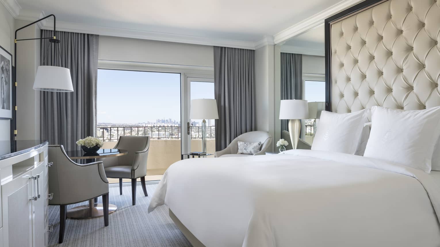 Premier Balcony Room hotel bed against padded, tufted wall, mirror, cafe table by sunny window