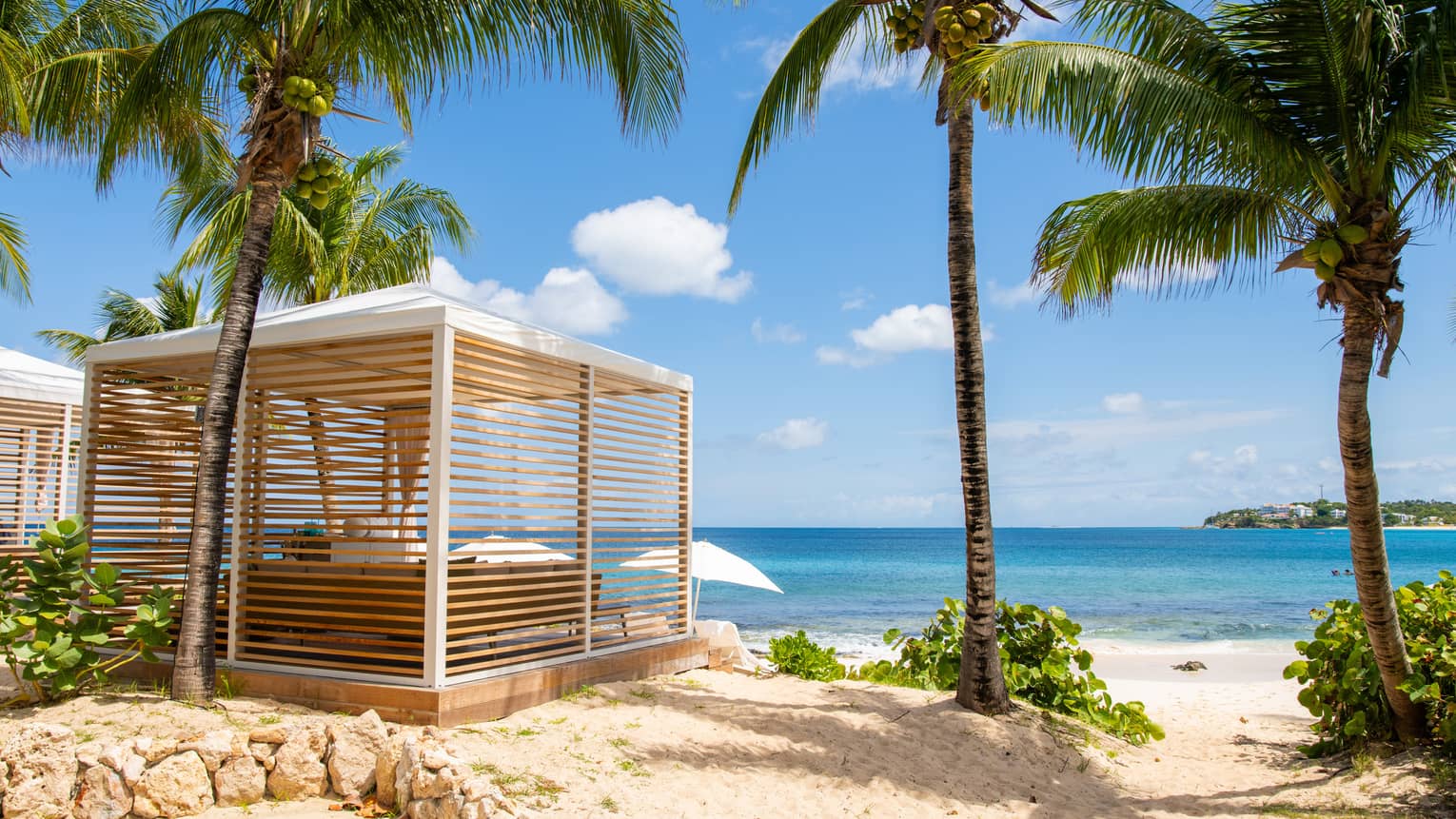 Luxury resort cabana with wooden slats surrounded by palm trees, located on a sandy beach and overlooking the ocean 