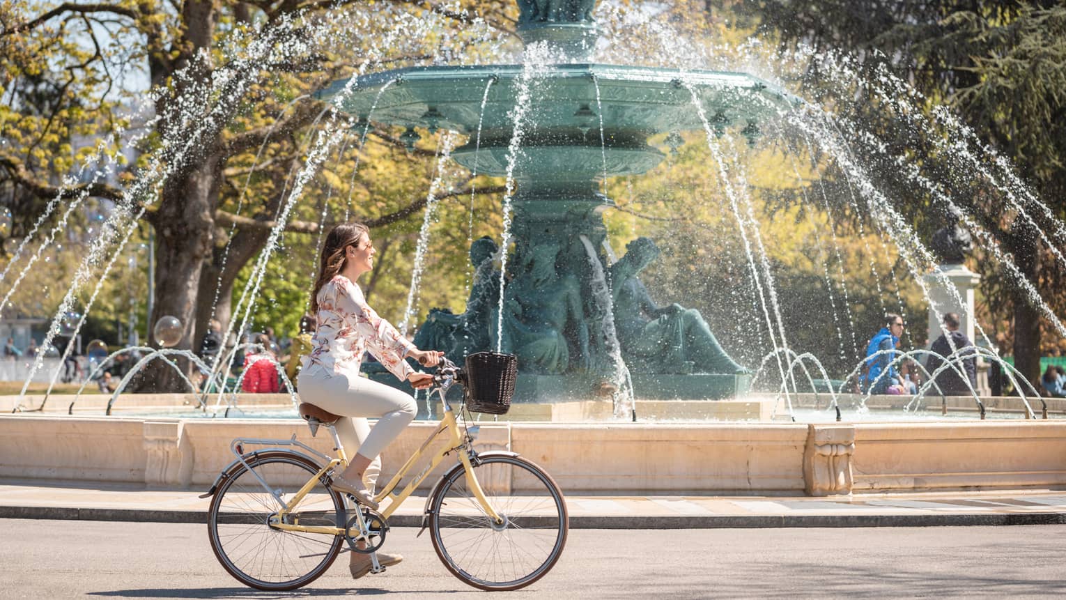 Side view of a guest on an old-style bike with a basket whizzing by a majestic fountain with a spout made from green statues.