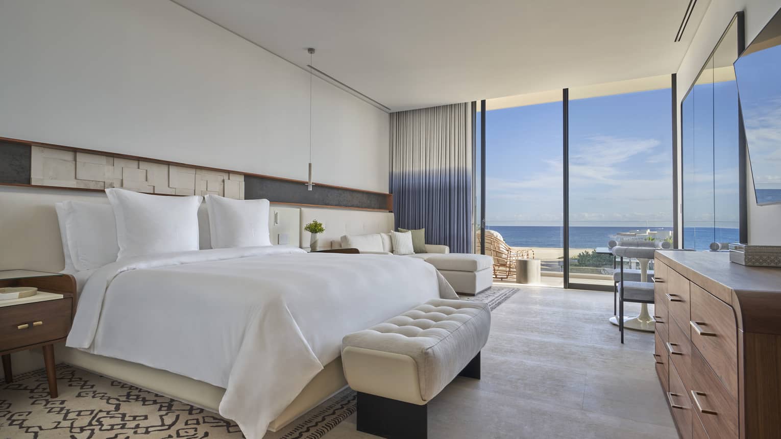 White bed with upholstered foot bench, side tables, dresser, lounge chair, tall glass doors and water views