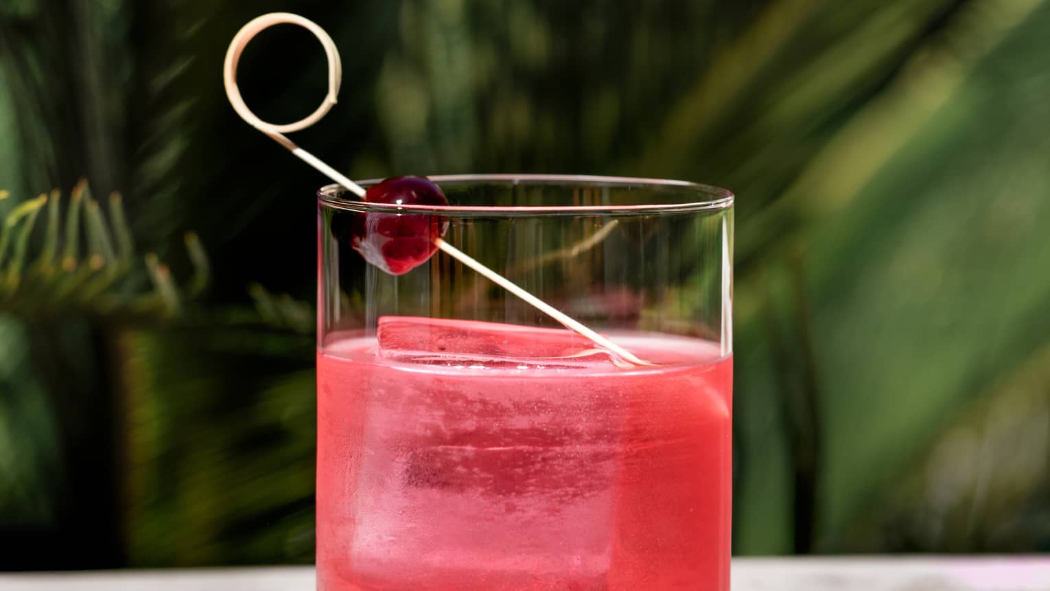 Red-hued cocktail in a rocks glass garnished with a large ice cube and a cherry on a curled toothpick