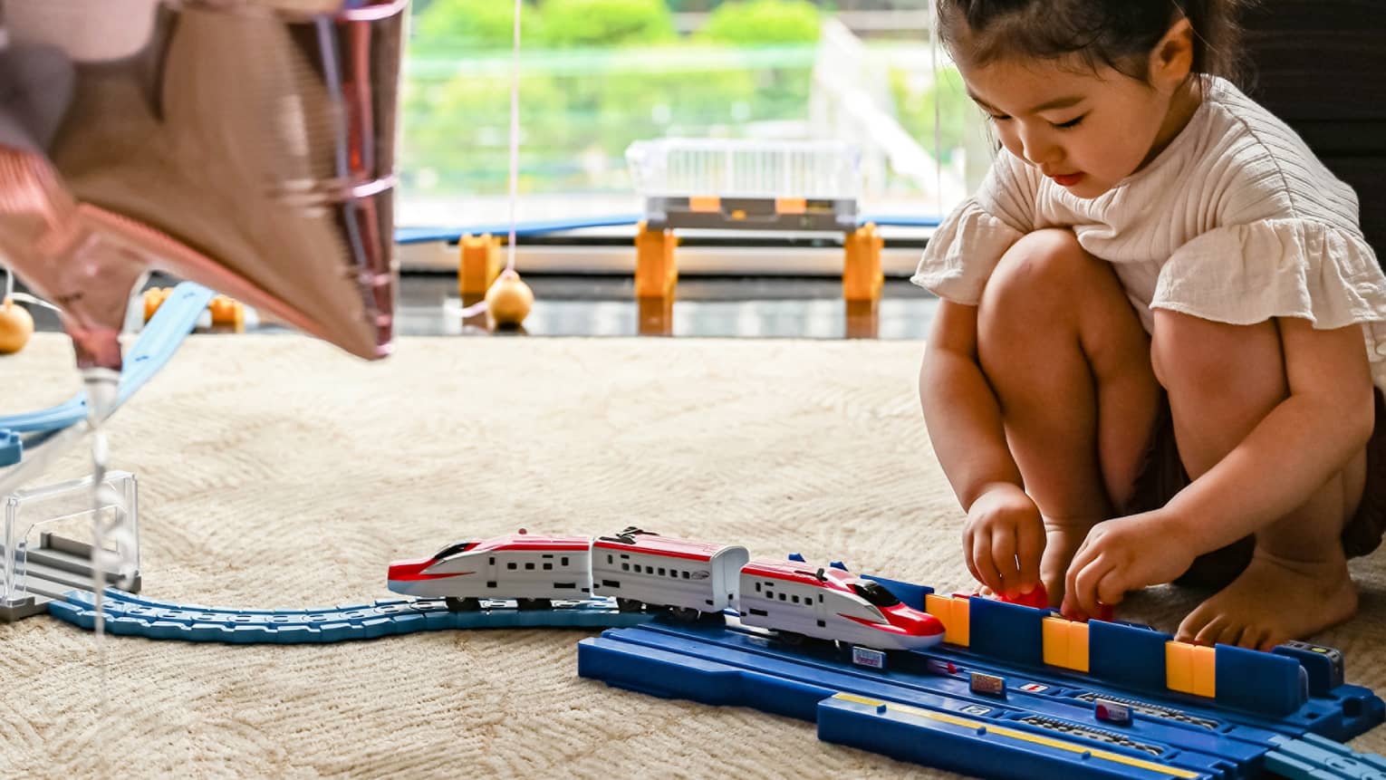 Young girl plays with train, large window and star balloons in the background
