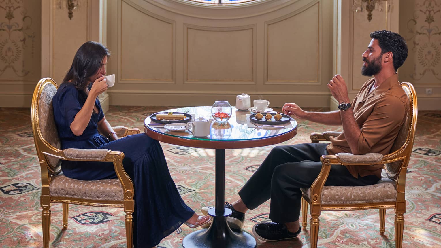 Couple sits across from each other sipping tea at small round table in front of large round window adorned with blues and greens