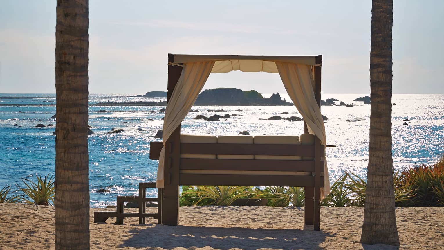 Steps lead up to a curtained daybed flanked by palm trees overlooking rocky islands in a sea of glimmering turquoise.