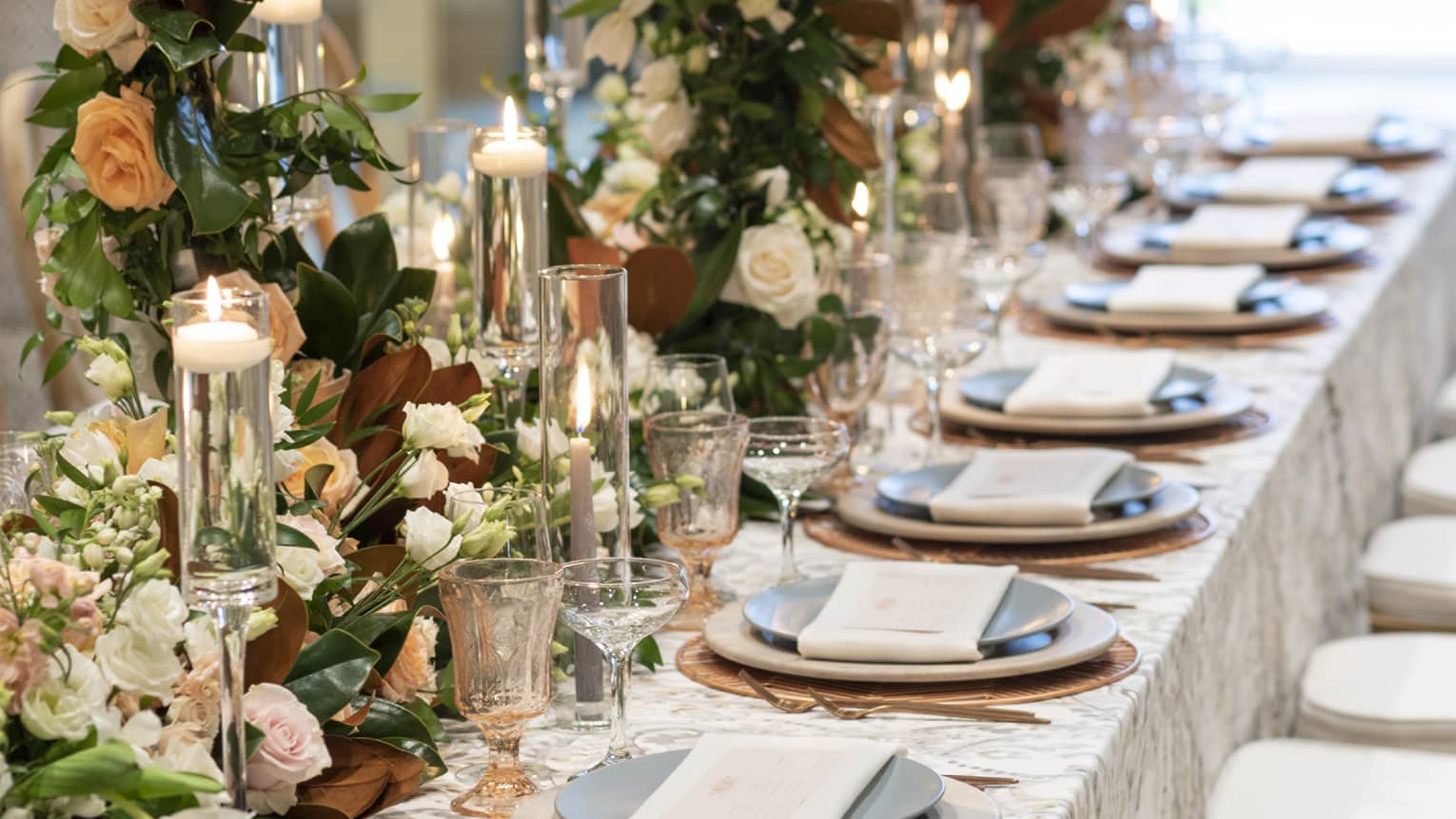 An elegant wedding reception table set at Four Seasons Resort Orlando, bathed in natural light from large windows. The table is adorned with a luxurious floral runner featuring ivory blooms and lush greenery, complemented by glowing candles in tall glass holders. Fine china, crystal glassware, and crisp linen napkins await guests, promising a sophisticated dining experience.