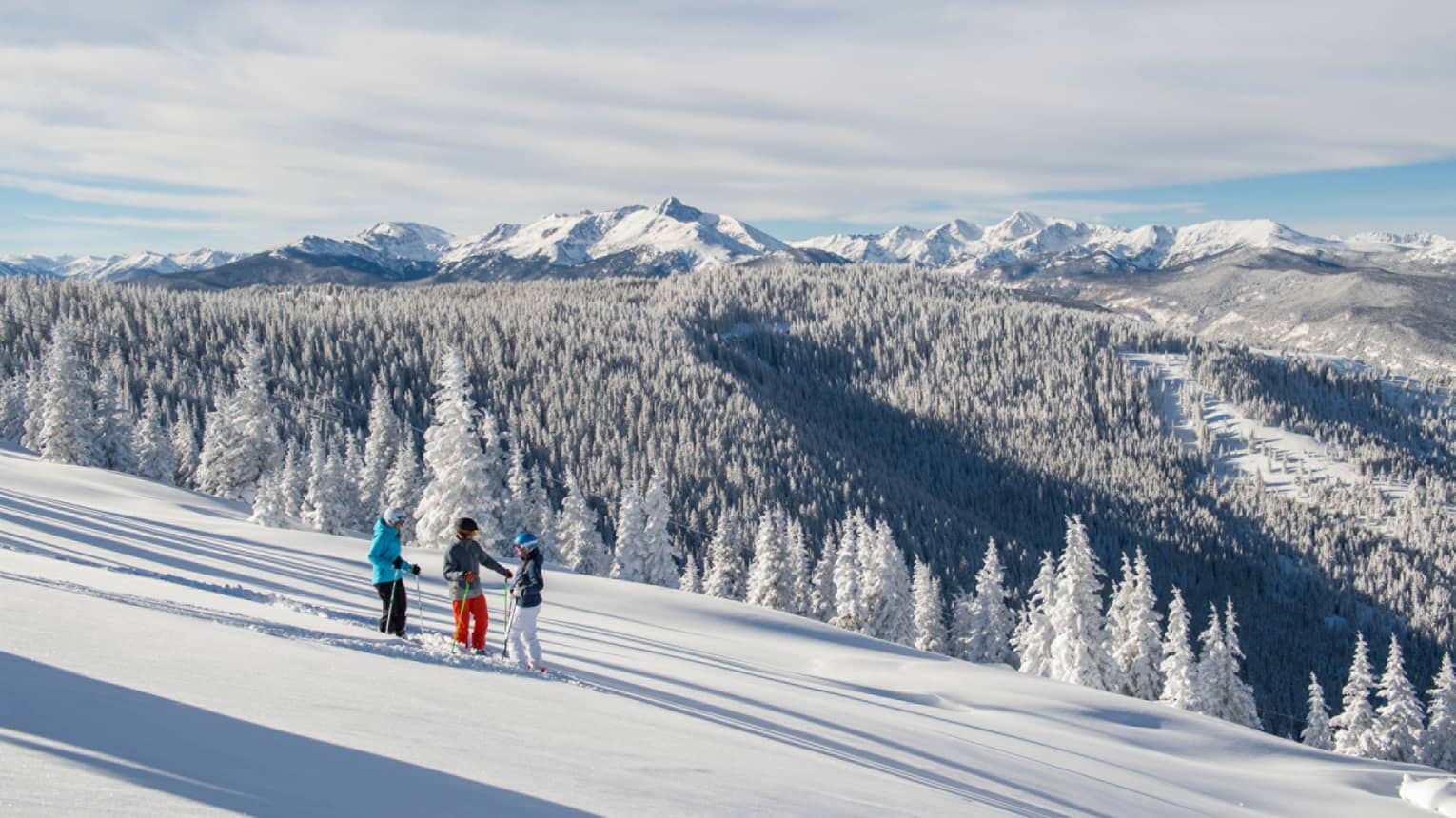 Three guests on skis pause on the ski hill, snow capped trees and mountains in distance 