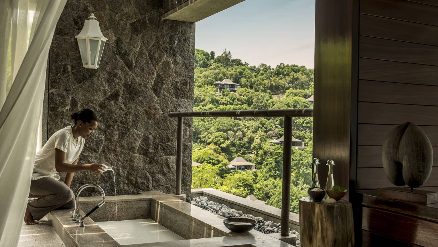 Woman kneels beside small rectangular spa tub, pours liquid into water, stone wall and trees