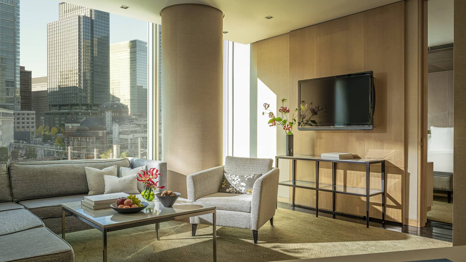 Four Seasons executive suite with gray sectional sofa, armchair, coffee table with fruit and flowers, large window with city view, TV and console table 