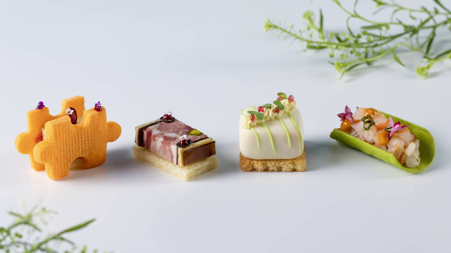 On a white table sit four small, savoury bites in various shapes, two of them on toast bases, with delicate floral accents.