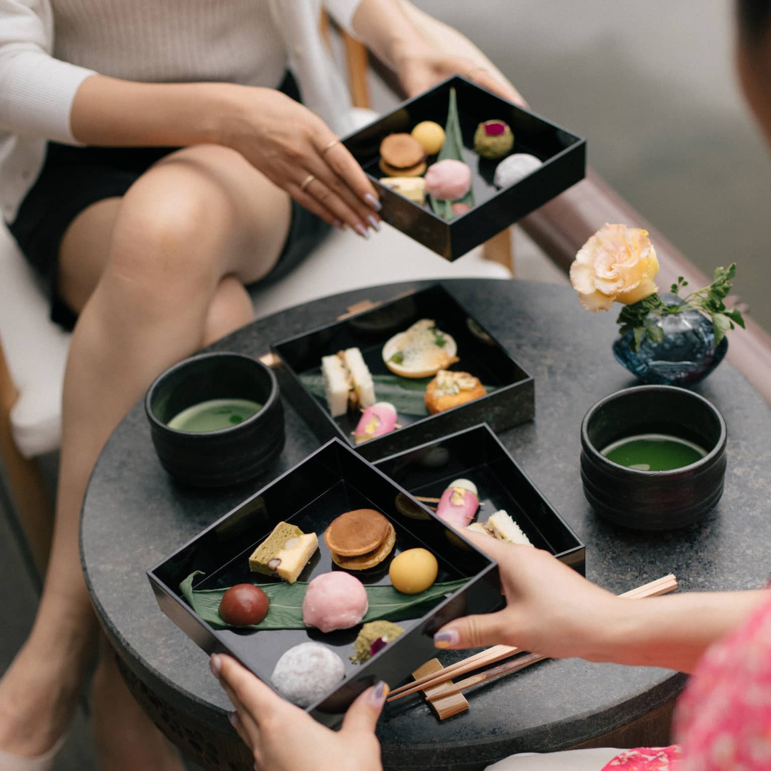 A small round table, with two-layered bento box of colourful, artfully arranged foods, top layers being lifted by two people.