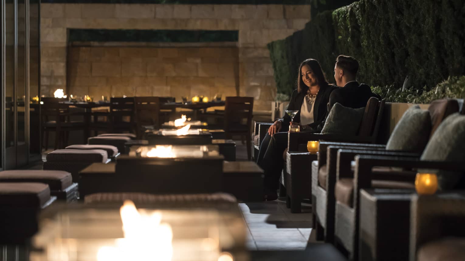 Two people in business attire sit in candle-lit cocktail lounge chairs with martinis