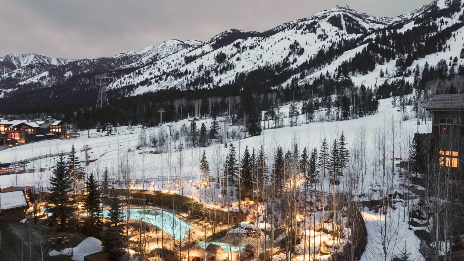 Aerial view of illuminated blue outdoor swimming pool through trees on snowy hill at dusk