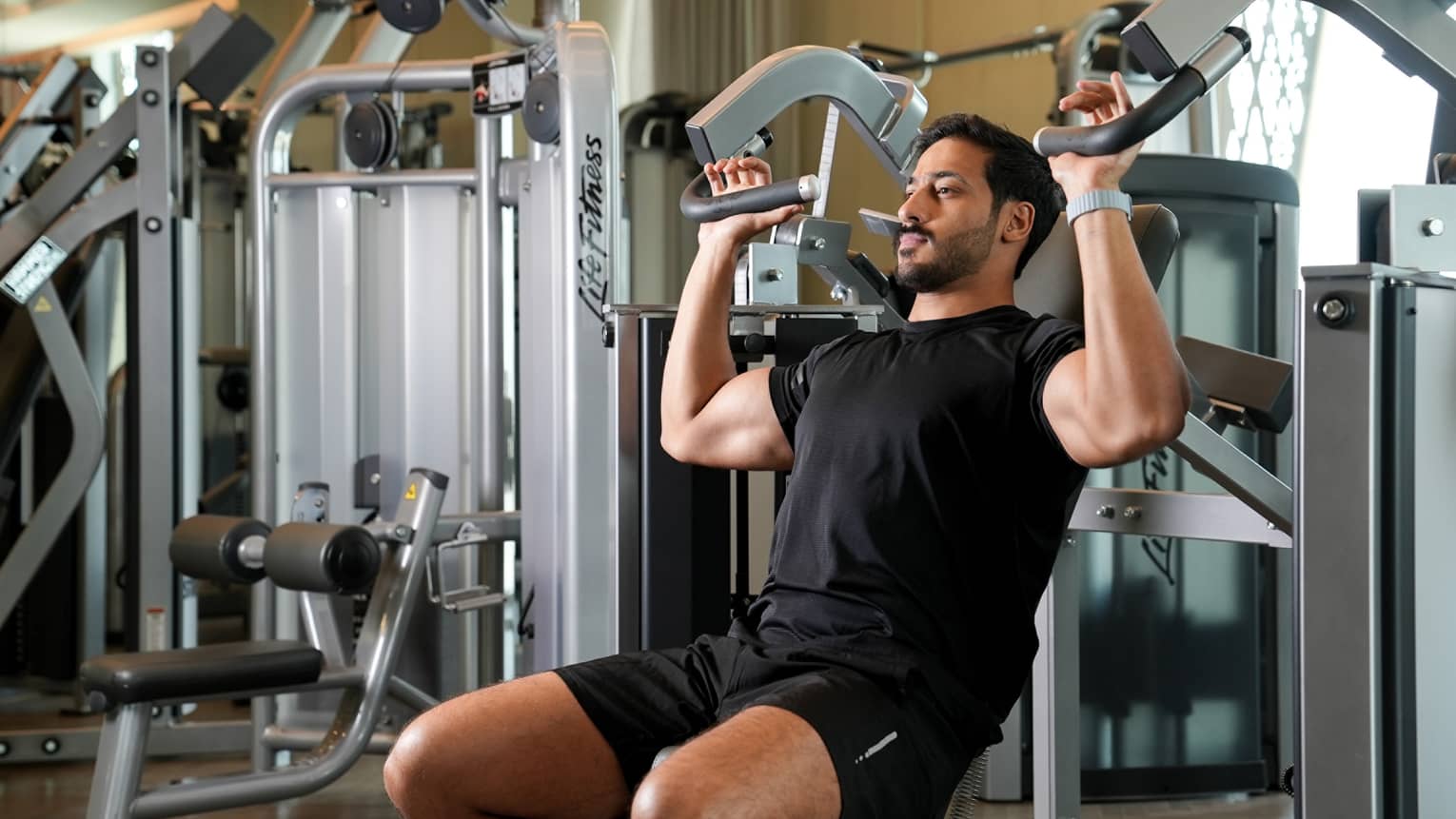 Man wearing black t-shirt and shorts works out on an arm weight machine at the gym