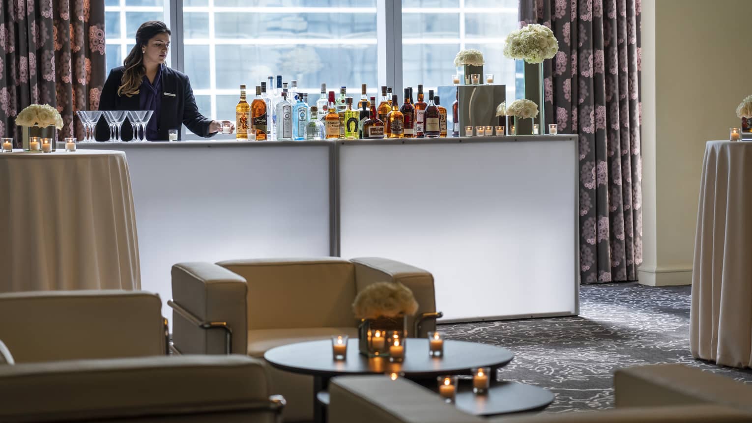 Bartender stands at liquor bottle-lined bar in front of window near seating area