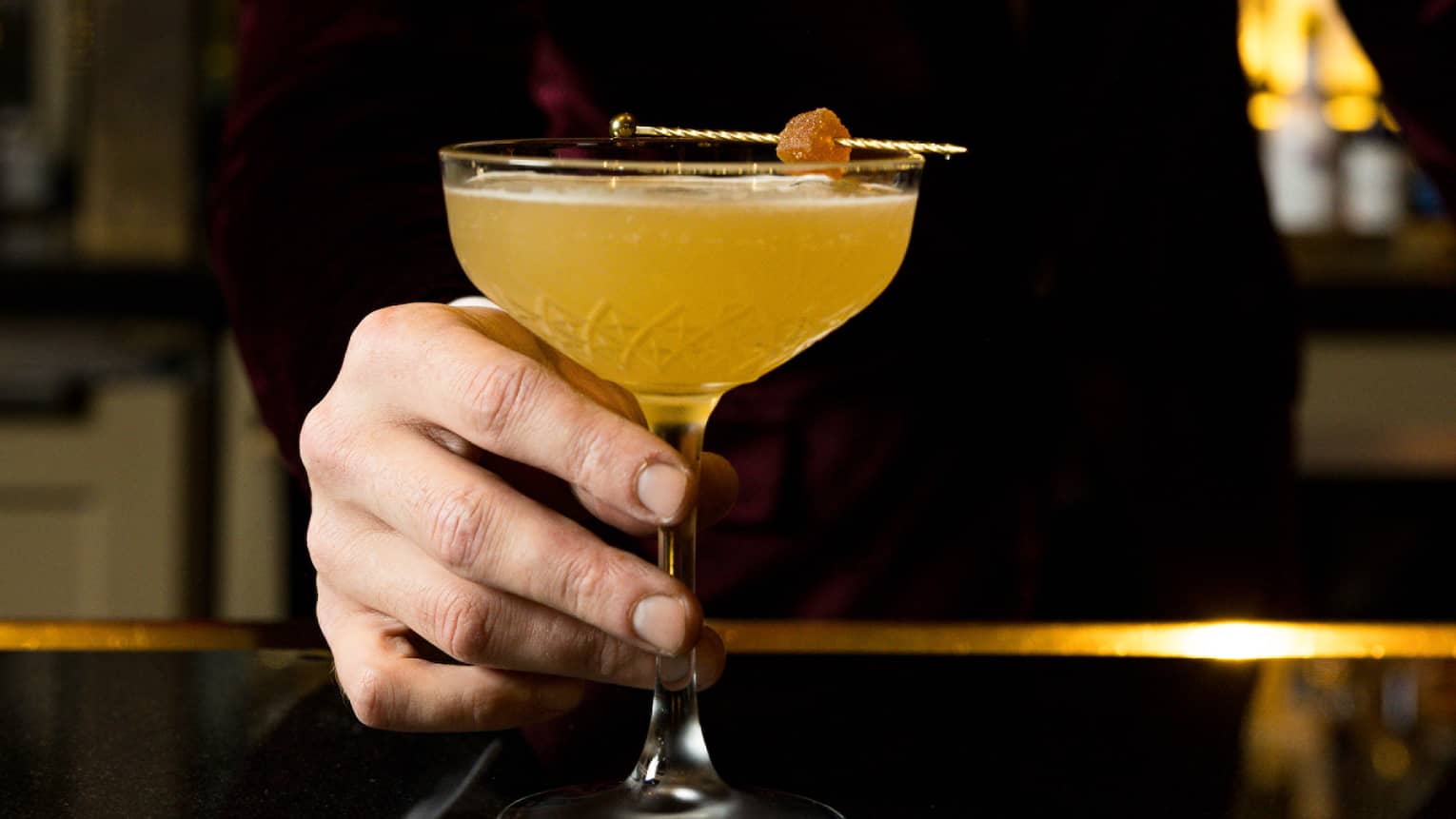A bartender dressed in a velvet tuxedo serves an orange cocktail at a bar