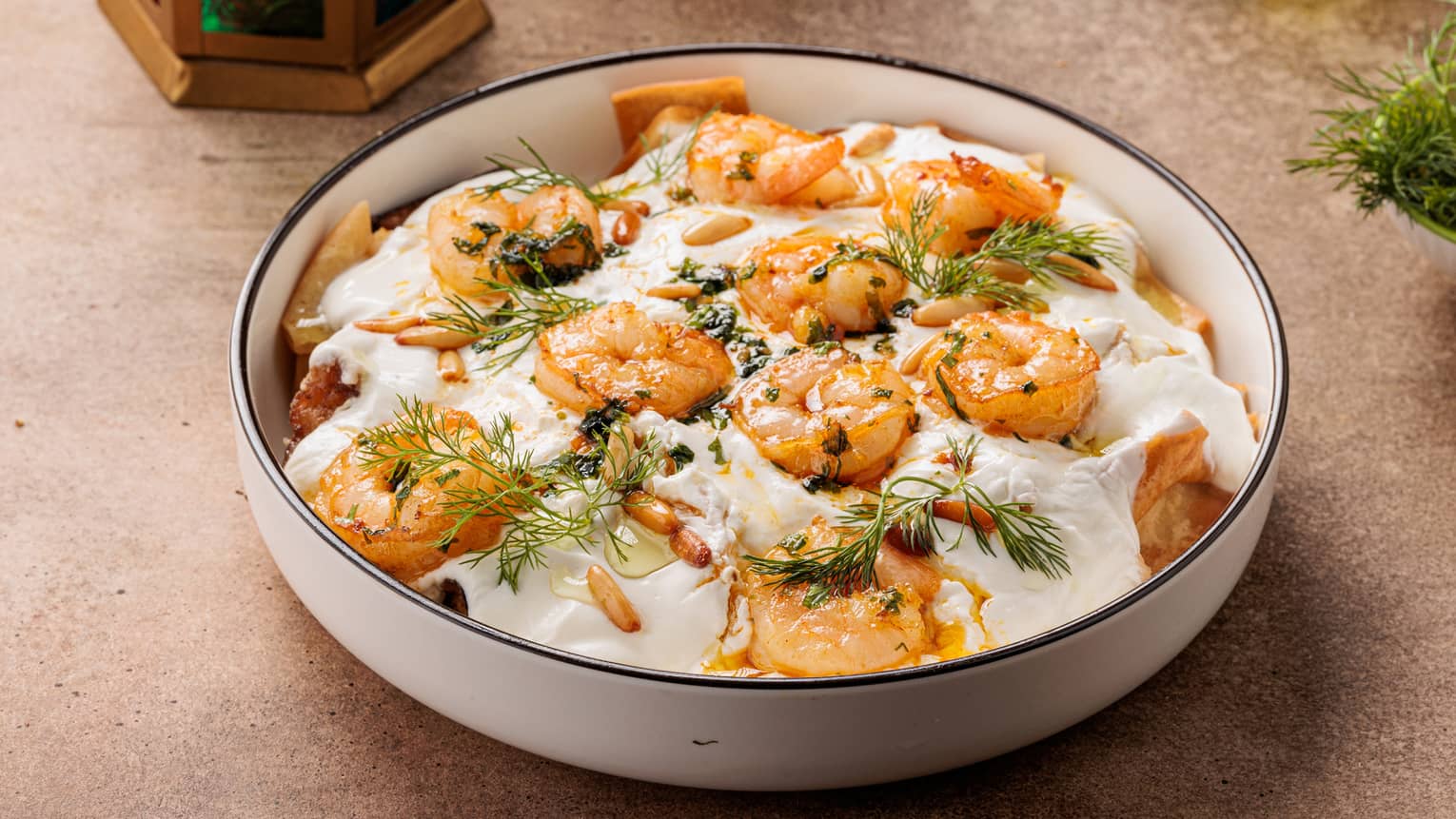 Shrimp in a white bowl served in a creamy yoghurt sauce next to a large jug of olive oil.