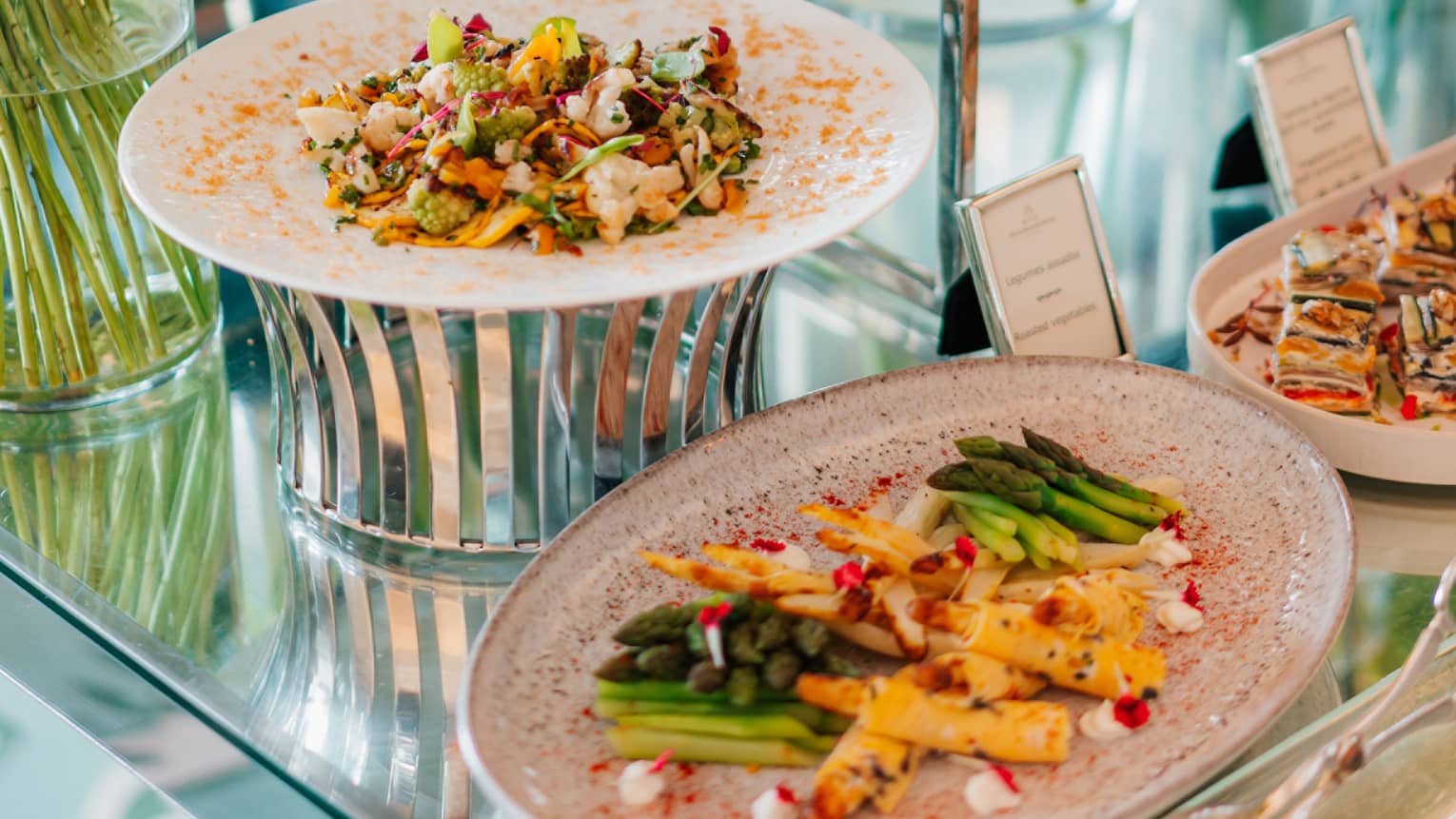 A table with a reflective surface holds vases of flowers and displays of colourful food items