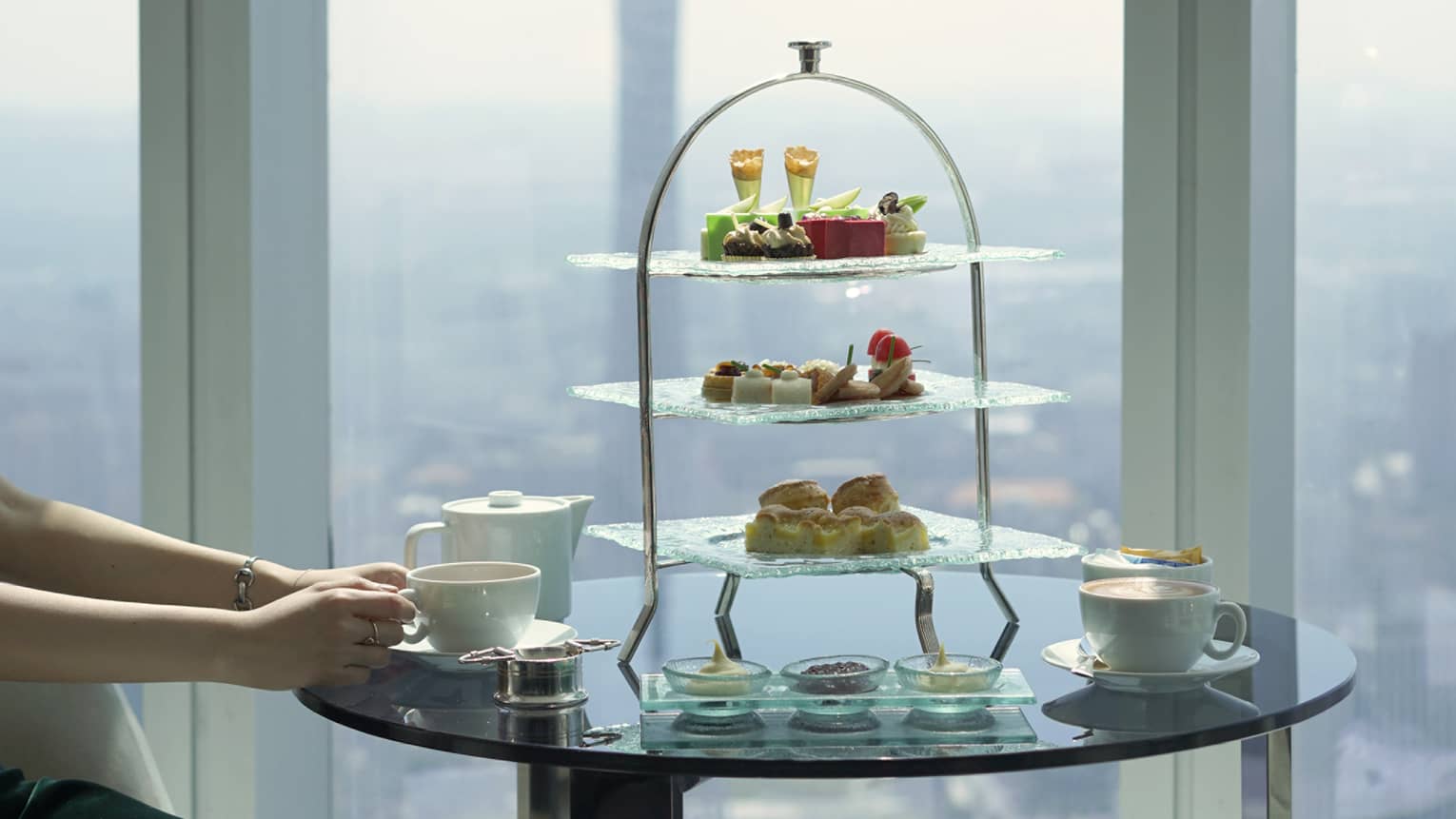 Close-up of a three-tiered tray and high tea setting on a glass table, a modern tower soaring skyward out the window beyond.