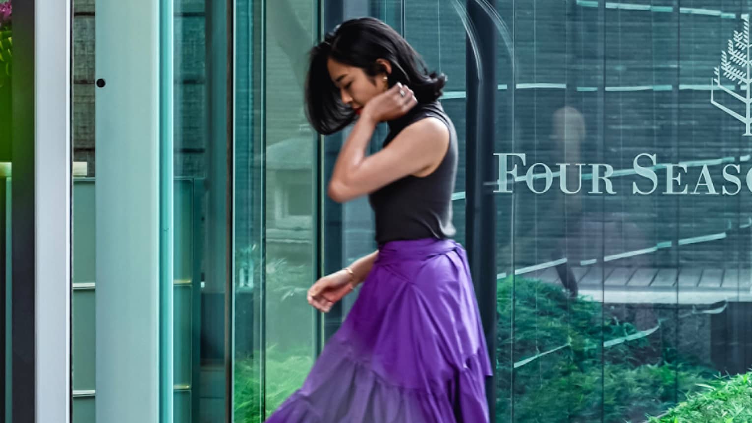 Person in a sleeveless top and purple skirt walking towards building with Four Seasons logo on glass door