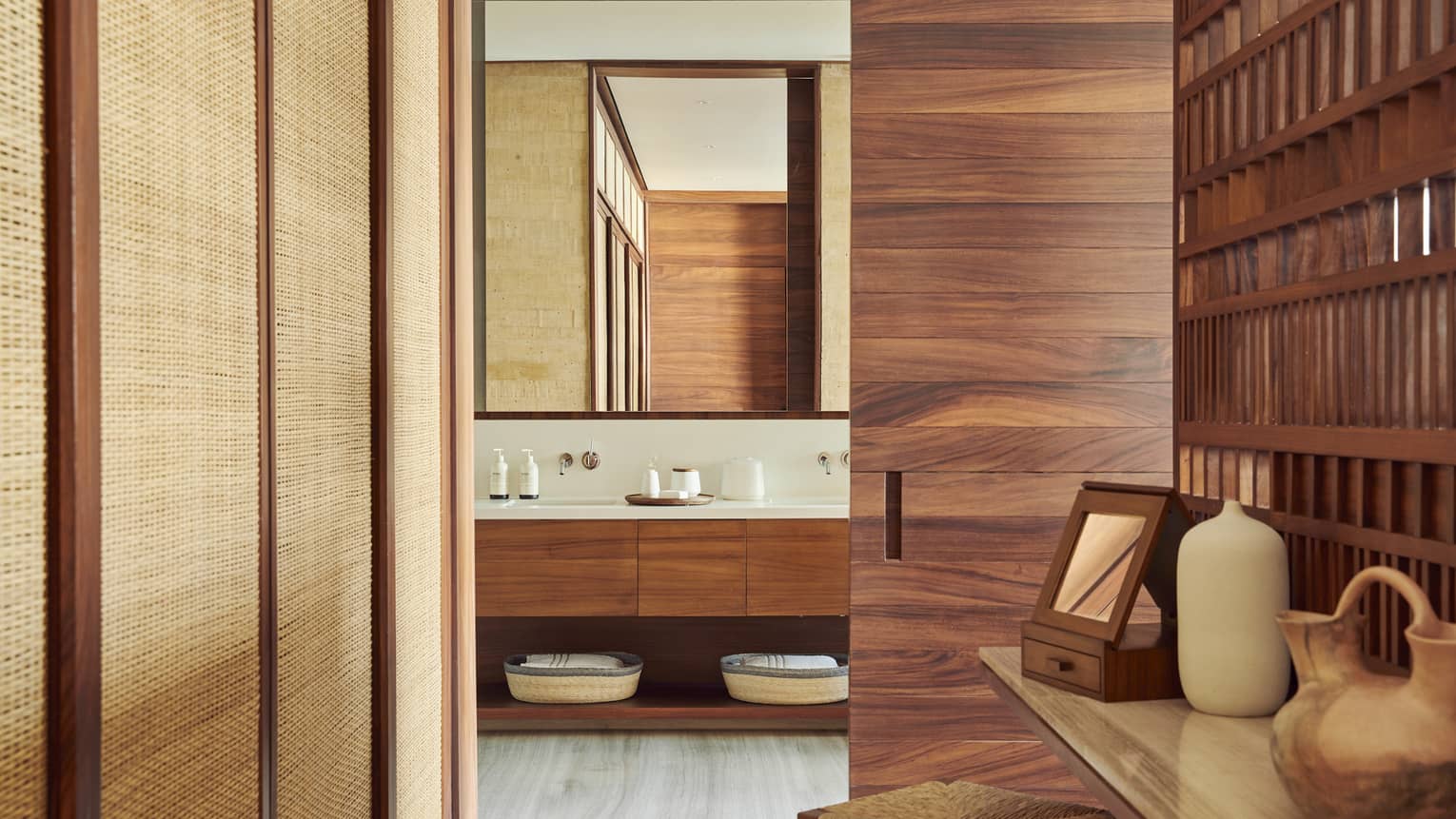 A modern bathroom entrance with wooden accents, featuring a double vanity with a large mirror. A nearby table holds decorative pottery and a small mirror.