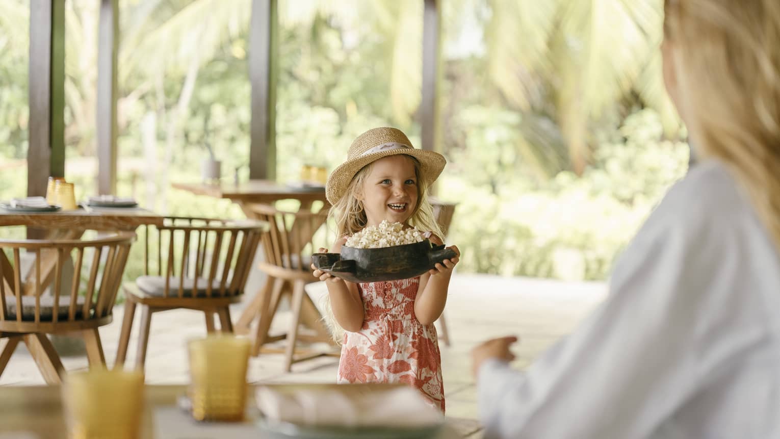 A small smiling child carrying a wooden turtle-shaped bowl filled with popcorn to their parent waiting on a patio.